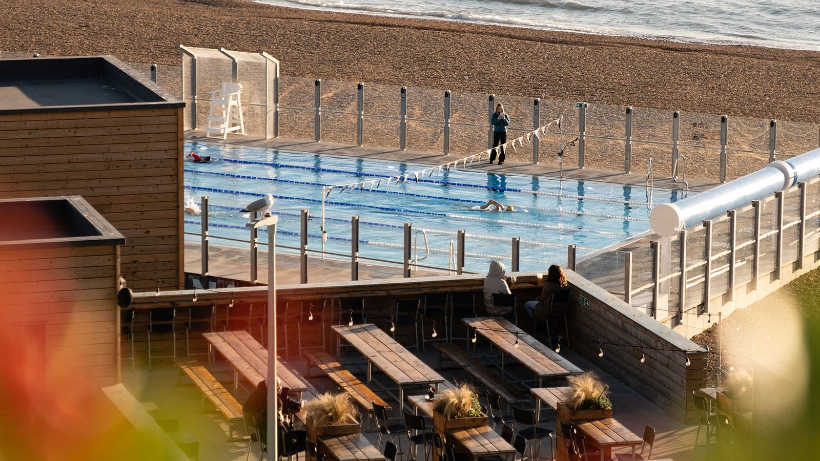 The Sea Lanes 50m outdoor pool on Brighton seafront at golden hour, with the beach and sea beyond and the deck terrace in the foreground.